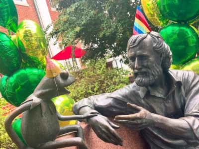 A photo of the Jim Henson statute with Birthday hats on Kermit and Jim Henson. There are green and gold balloons behind the bench.