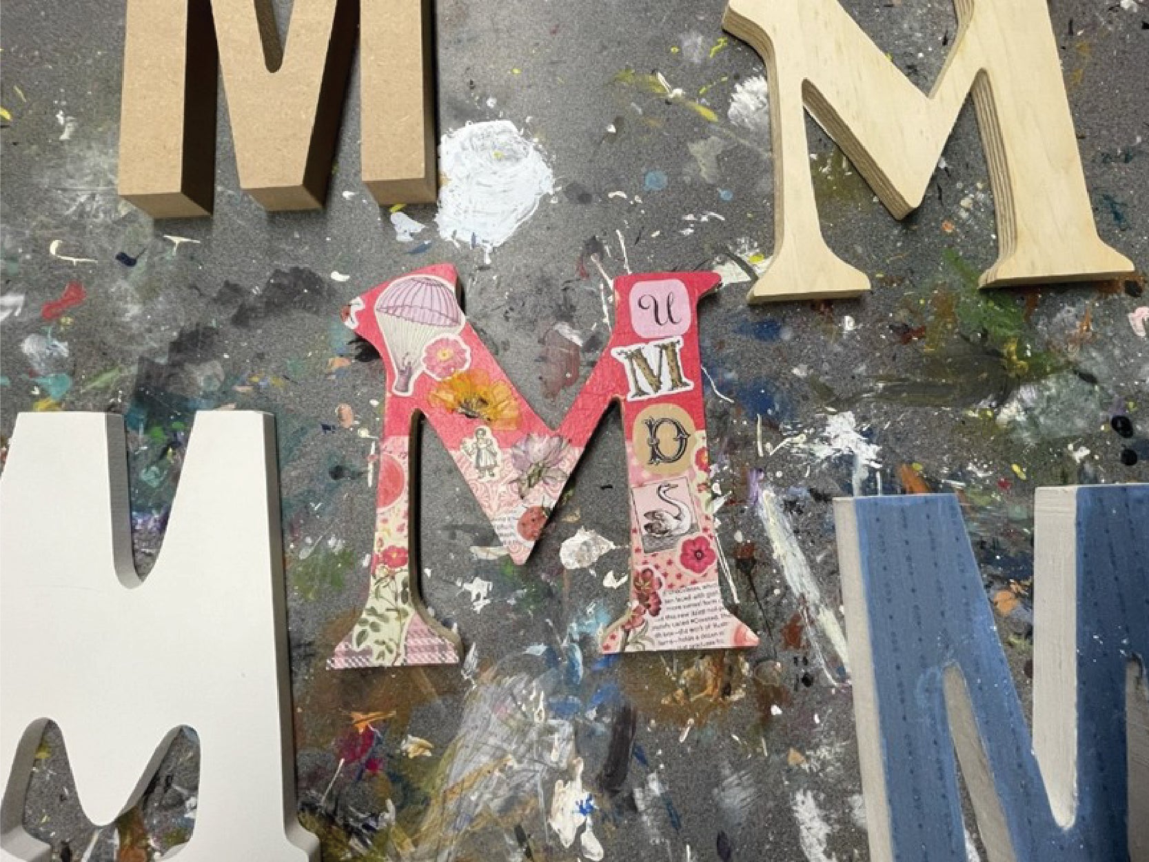 Five wooden "M"s, one of which is decoupaged with a variety of whimsical collage elements