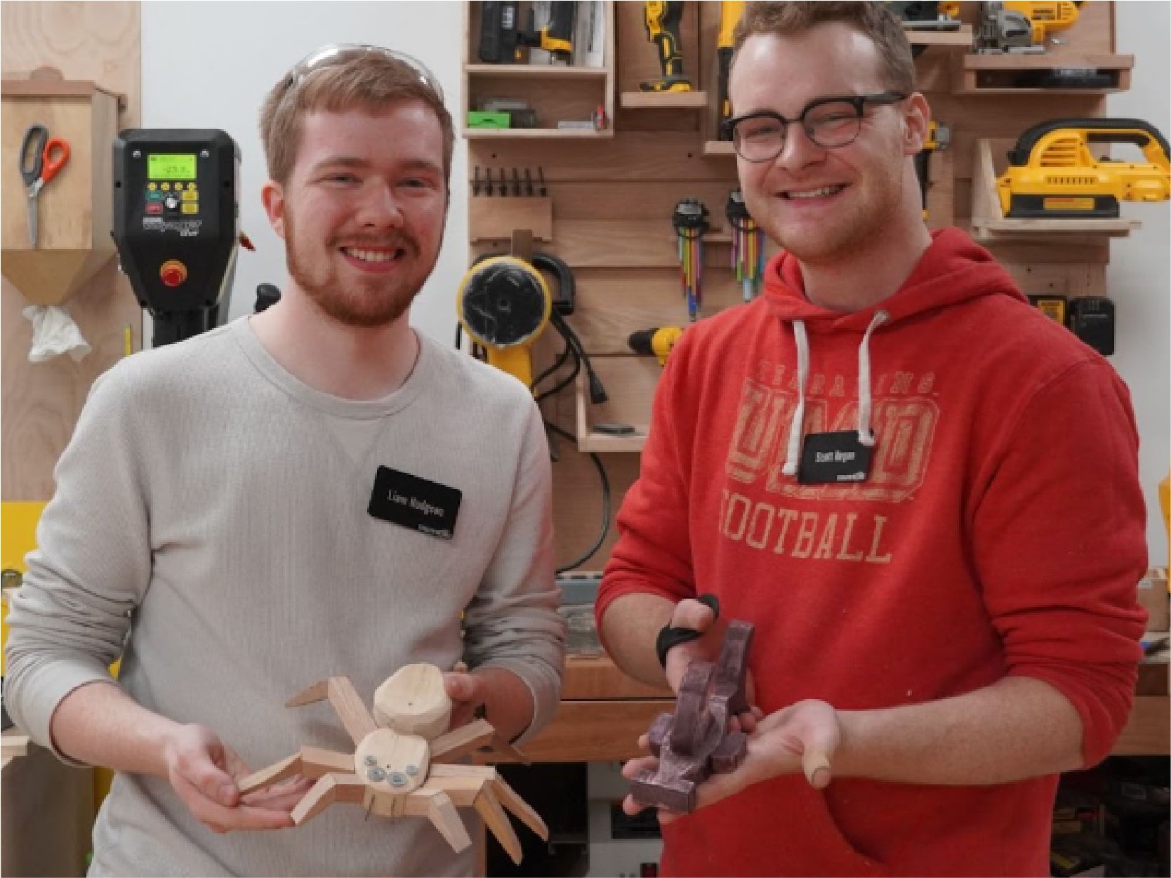 Two men stand in a woodworking shop holding a spider and a shark crafted from wood.