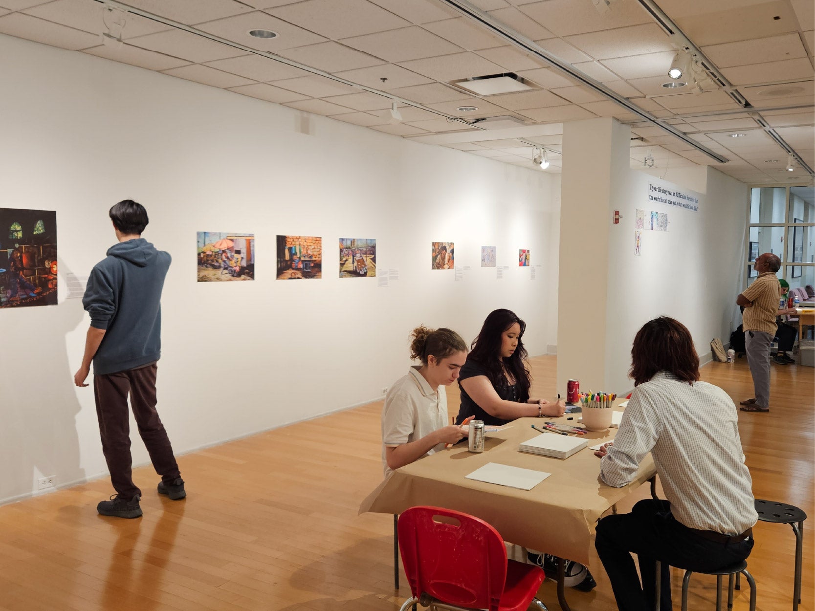 Visitors in the STAMP Gallery engage with artwork and creative activities. Two visitors stand looking at artwork, while three others sit at a table drawing.