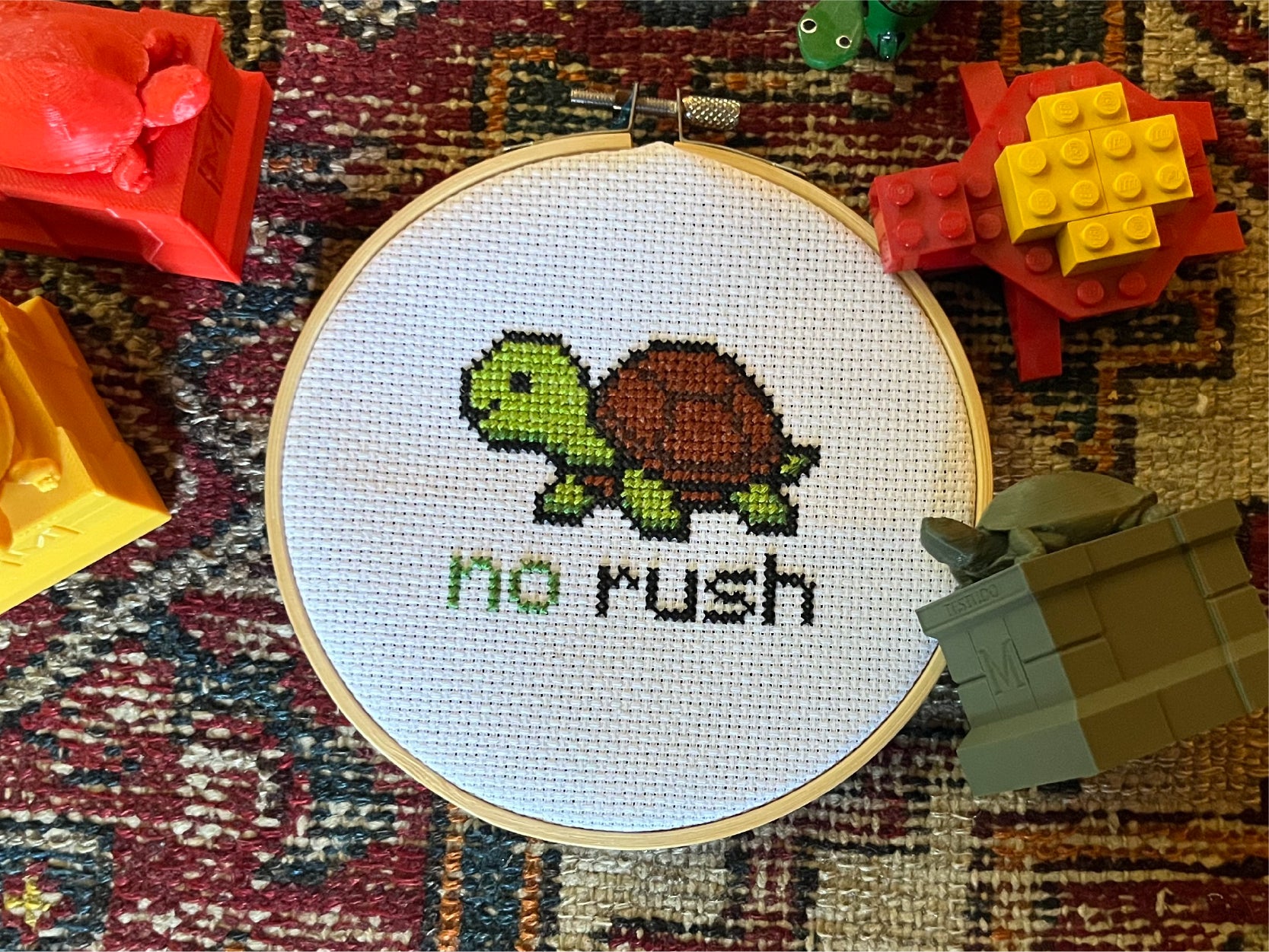 A photo of a testudo cross-stitch project surrounded by 4 mini testudo toys in various colors.