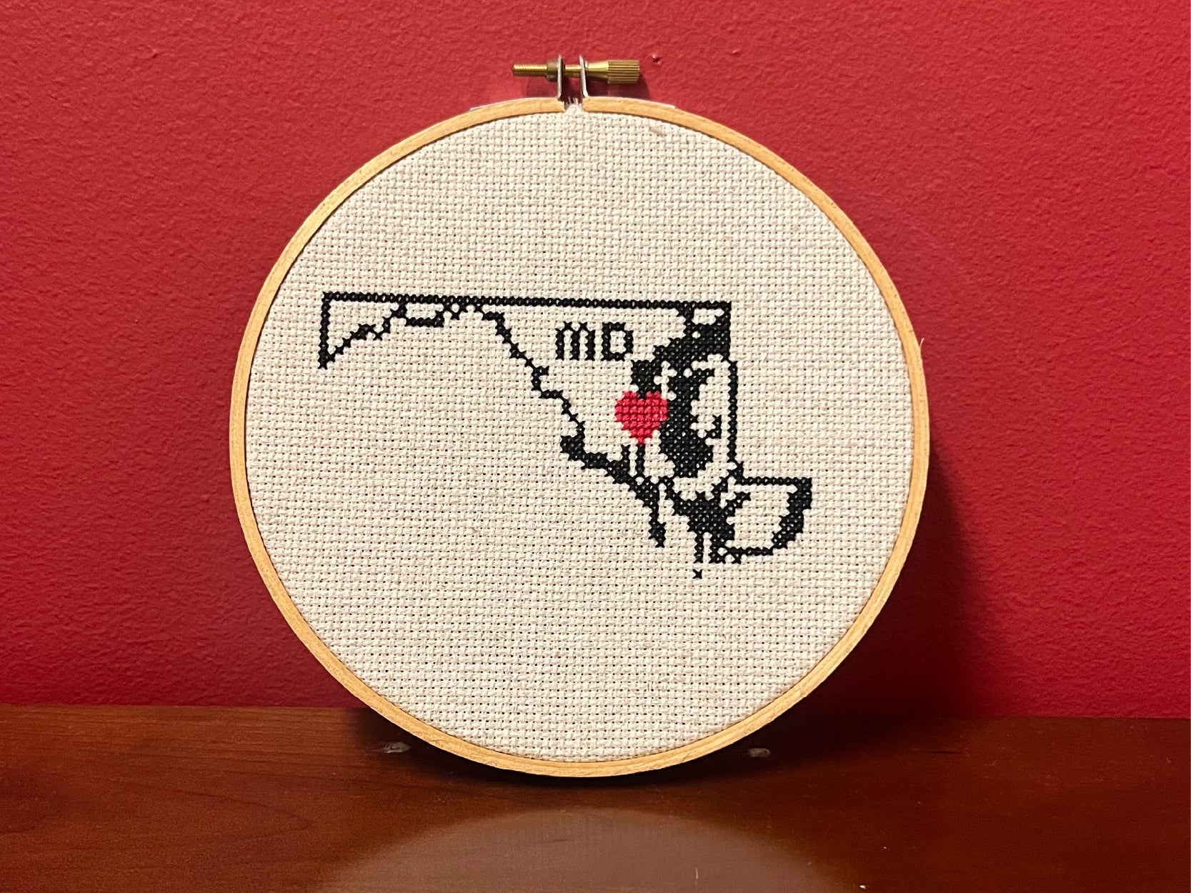 A photo of a cross-stitch project. It is a black outline of the State of Maryland with a red heart in the middle.